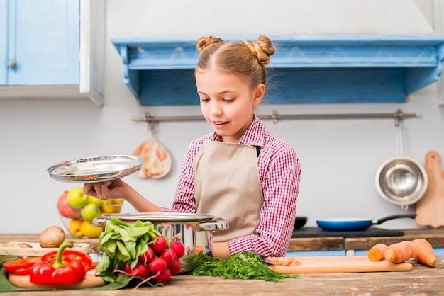 Retrato de una niña sonriente mirando acero inoxidable cocina olla en la cocina