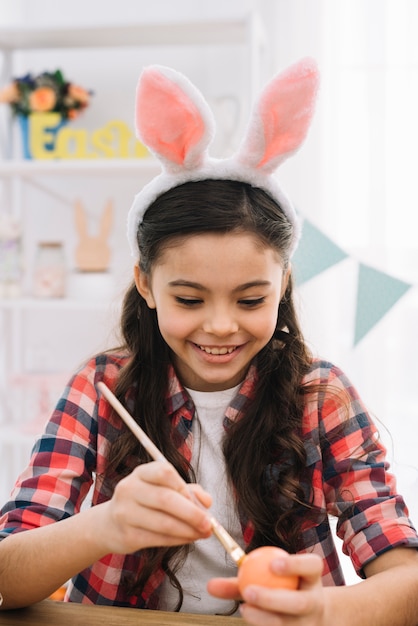 Retrato de una niña sonriente con orejas de conejo pintando huevos de Pascua con pincel