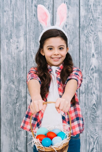 Retrato de una niña sonriente con orejas de conejo que ofrece la canasta de huevos de Pascua