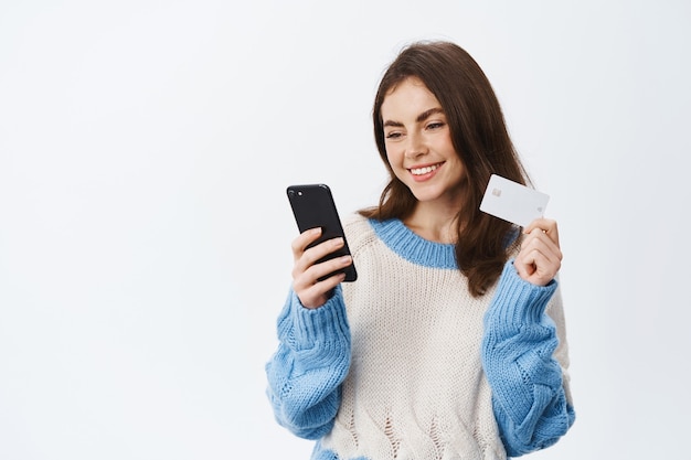 Retrato de niña sonriente pagando en la tienda de internet con tarjeta de crédito plástica y aplicación móvil, sosteniendo el teléfono inteligente y leyendo la pantalla del teléfono, pared blanca