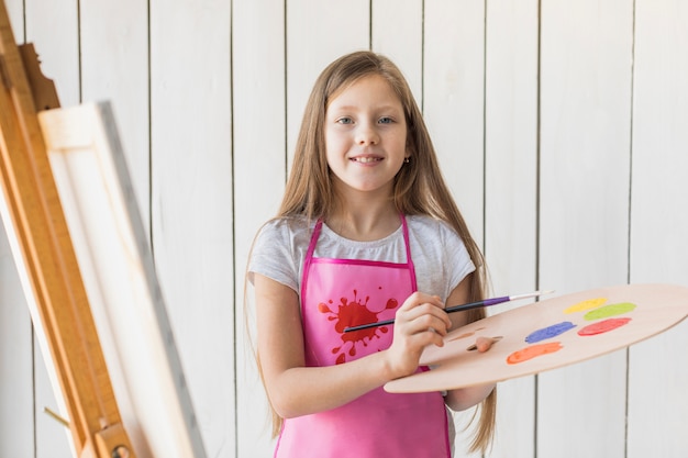 Retrato de una niña sonriente con paleta de madera y pincel de pie contra la pared de madera blanca