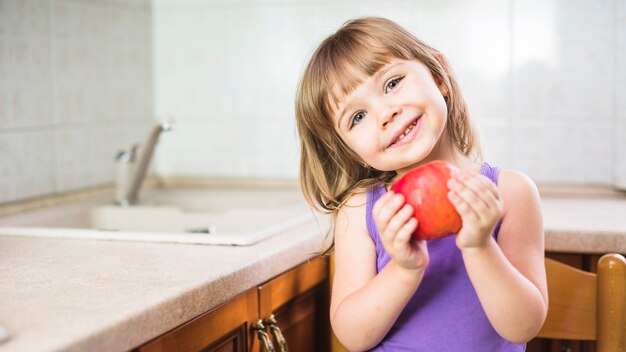 Retrato de una niña sonriente de pie en la cocina con manzana roja fresca