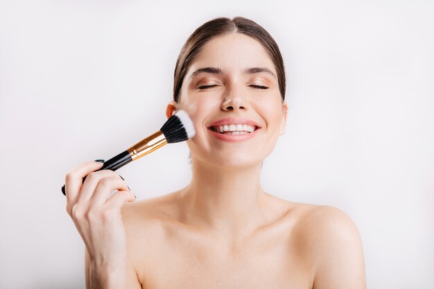 Retrato de niña sonriente con piel limpia aplicando pincel para polvos en la cara contra la pared aislada.