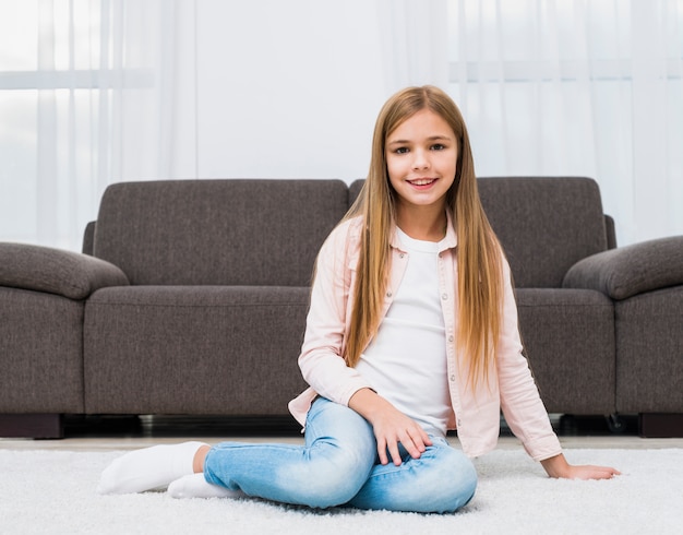 Retrato de niña sonriente sentada en la alfombra delante del sofá mirando a la cámara