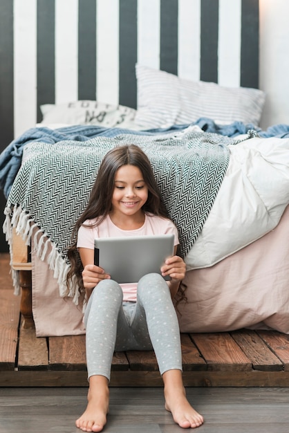 Retrato de una niña sonriente sentada cerca de la cama mirando tableta digital