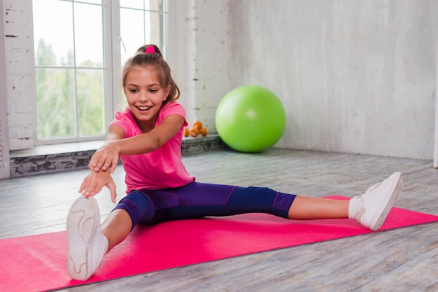 Retrato de una niña sonriente sentada en una estera de ejercicios estirando su mano y pierna