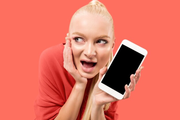 Retrato de una niña sorprendida, sonriente, feliz y asombrada que muestra el teléfono móvil de pantalla en blanco aislado sobre fondo coral.