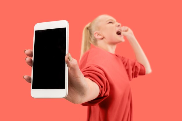 Retrato de una niña sorprendida, sonriente, feliz y asombrada que muestra el teléfono móvil de pantalla en blanco aislado sobre fondo coral.