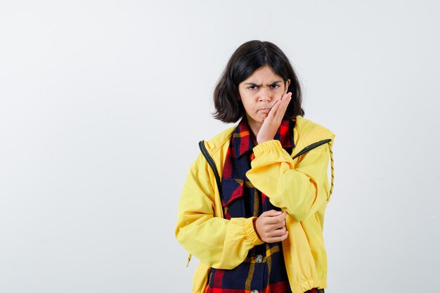 Retrato de niña sosteniendo la mano en la mejilla en camisa a cuadros, chaqueta y mirando enojado vista frontal