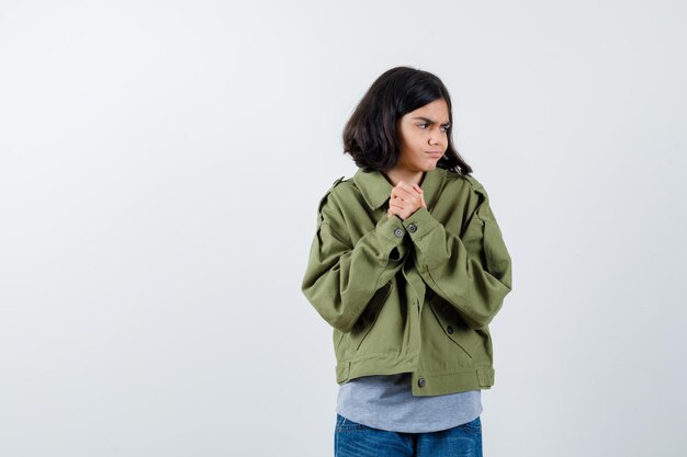 Retrato de niña sosteniendo las manos juntas mientras mira a otro lado en abrigo, camiseta, jeans y mirando pensativa vista frontal