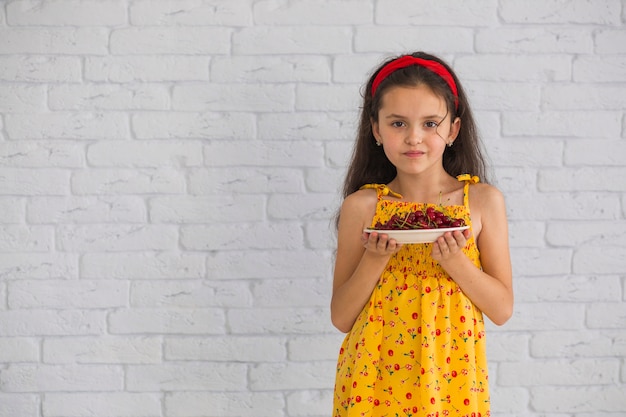 Retrato de niña sosteniendo la placa de cerezas rojas contra la pared de ladrillo blanco