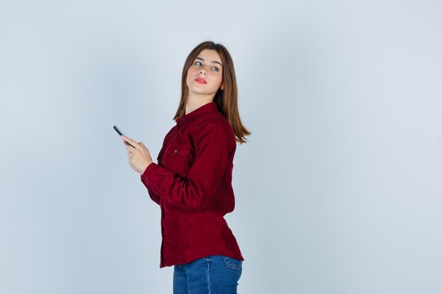 Retrato de niña sosteniendo el teléfono móvil mientras mira hacia atrás en camisa burdeos y mirando curioso