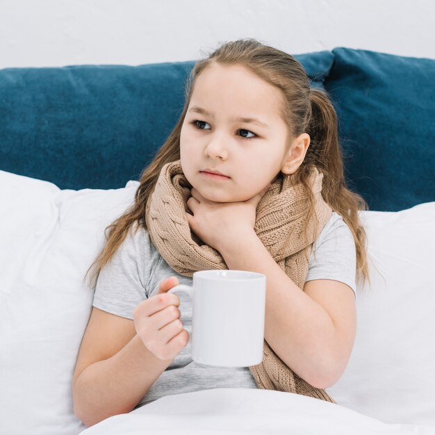 Retrato de una niña tocando su garganta sosteniendo una taza de café en la mano