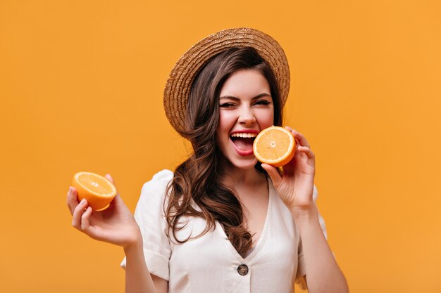Retrato de niña traviesa con cabello ondulado mordiendo naranja. Señora con sombrero de paja posando sobre fondo naranja.