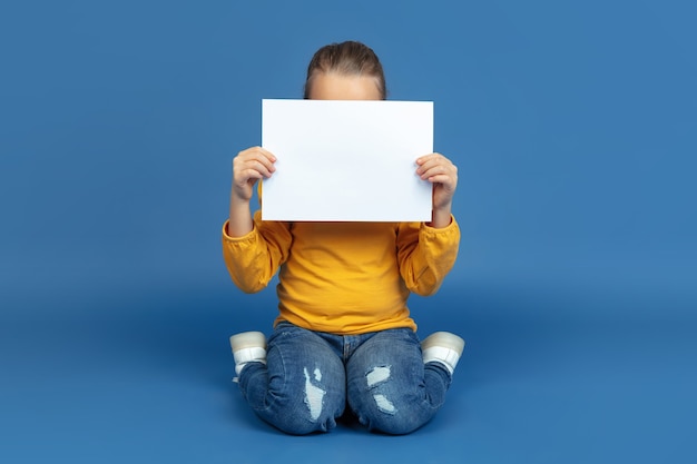 Retrato de niña triste sentada aislada sobre fondo azul de estudio.