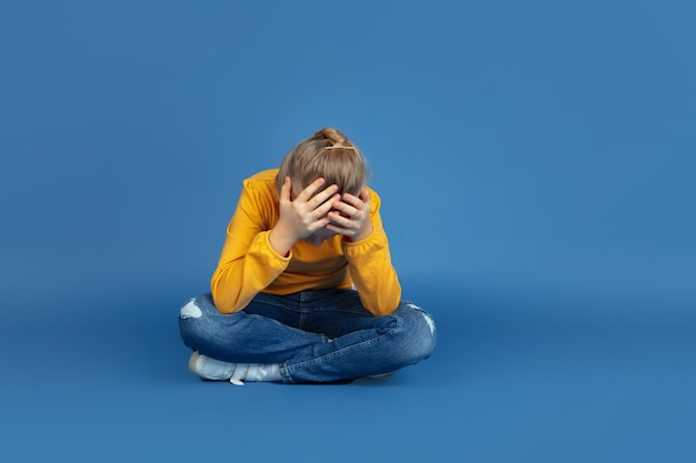 Retrato de niña triste sentada aislada sobre fondo azul.