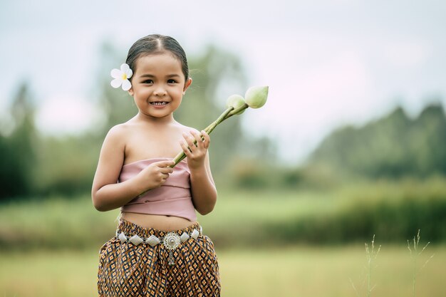 Retrato de niñas encantadoras en traje tradicional tailandés y poner flor blanca en su oreja, de pie y sostener dos loto en la mano en el campo de arroz, sonríe con dientes, copia espacio