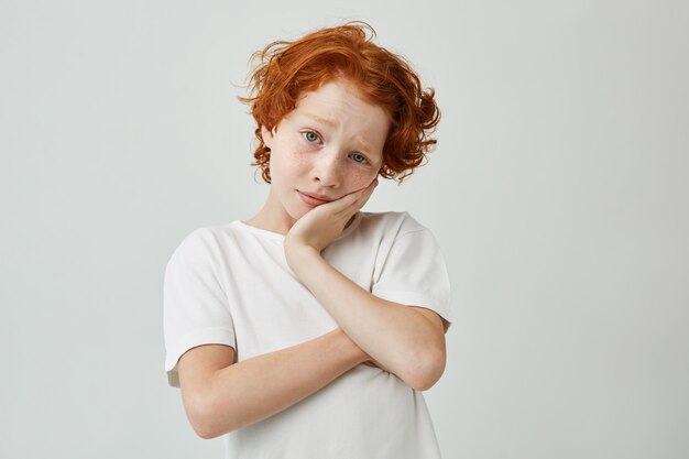 Retrato de niño bonito con pelo de jengibre y pecas sosteniendo la cabeza con las manos aburridas