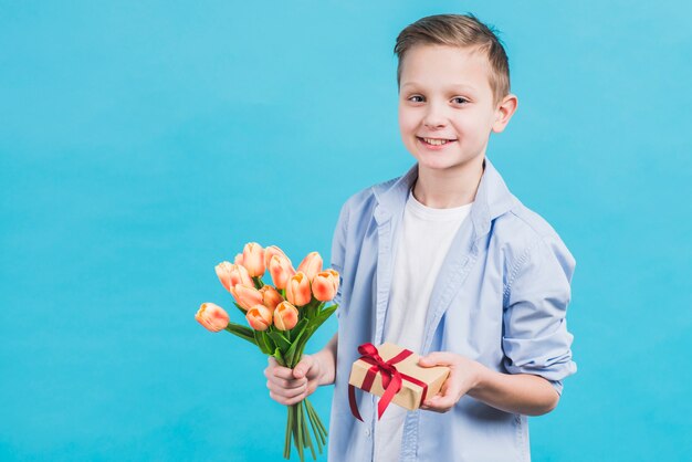 Retrato de un niño con caja de regalo envuelta y tulipanes en la mano contra el fondo azul