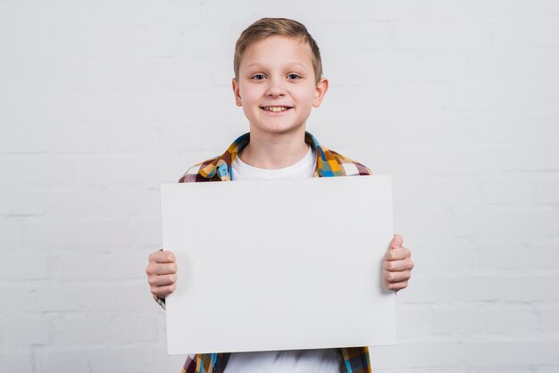 Retrato de un niño feliz de pie contra la pared blanca que muestra el cartel en blanco blanco