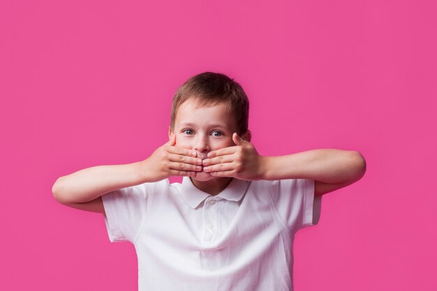 Retrato de niño inocente cubriendo su boca y mirando a la cámara sobre fondo de pared rosa