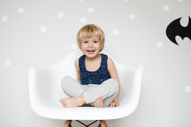 Retrato de niño juguetón con cabello rubio en pijama, sentado en una silla con las piernas cruzadas, riendo, con la boca abierta, mostrando sus dientes blancos, contra la pared en blanco ganar