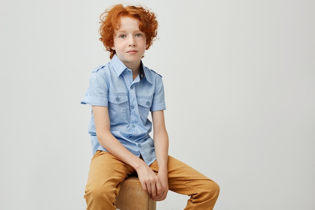 Retrato de niño lindo con cabello rizado jengibre mirando con expresión seria, sentado en una caja