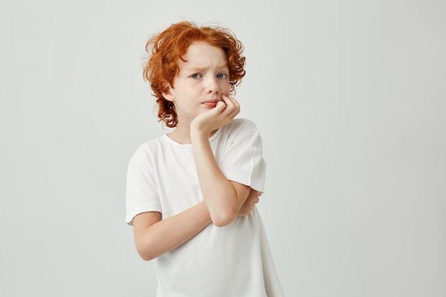 Retrato de niño lindo con cabello rojo y pecas sosteniendo la cabeza con la mano pensando en la tarea que debe hacer hasta la noche.