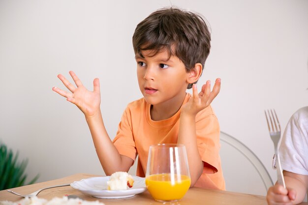 Retrato de niño lindo comiendo pastel de cumpleaños y bebiendo jugo de naranja. Adorable niño sentado a la mesa en el comedor, levantando las manos y mirando a otro lado. Concepto de infancia, celebración y vacaciones.