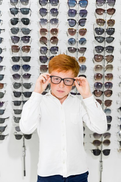 Retrato de niño lindo con gafas de pie contra el estante de las lentes en la tienda de óptica