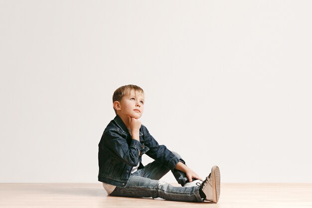 Retrato de niño lindo niño en ropa de jeans con estilo mirando a cámara contra la pared blanca del estudio. Concepto de moda infantil