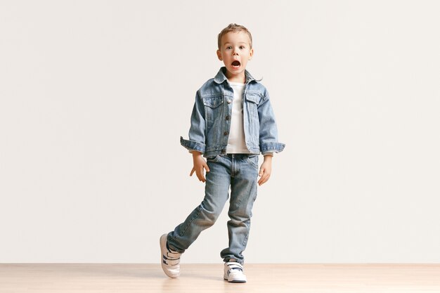 Retrato de niño lindo niño en ropa de jeans con estilo mirando a cámara contra la pared blanca del estudio. Concepto de moda infantil