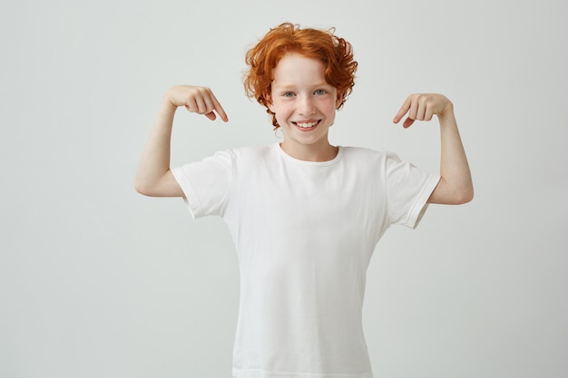 Foto gratuita retrato de niño lindo con pelo de jengibre apuntando con los dedos de ambas manos en camiseta blanca y sonriendo