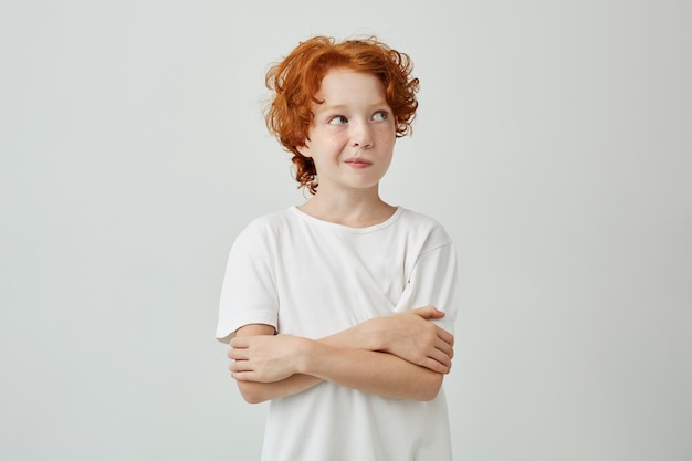 Foto gratuita retrato de niño lindo con pelo de jengibre y pecas en camiseta blanca mirando a otro lado, persiguiendo sus labios, con las manos cruzadas.