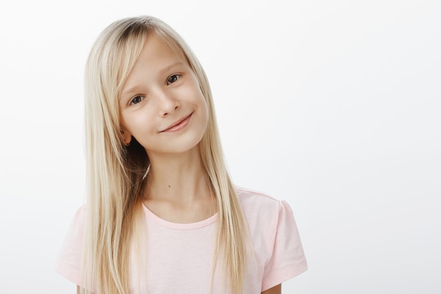 Retrato de niño lindo positivo complacido con sonrisa de satisfacción feliz, inclinando la cabeza mientras escucha con atención, siendo un buen niño obediente, de pie sobre una pared gris