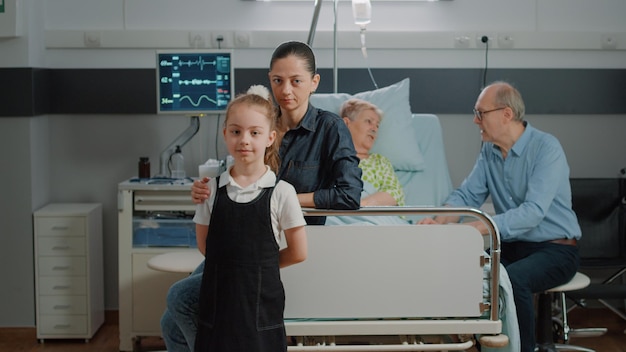 Retrato de niño y madre visitando a un paciente mayor en la cama de la sala del hospital. Mujer y niño sentados en la sala de cuidados intensivos en visita familiar, brindando apoyo al pensionista con enfermedad en la clínica.