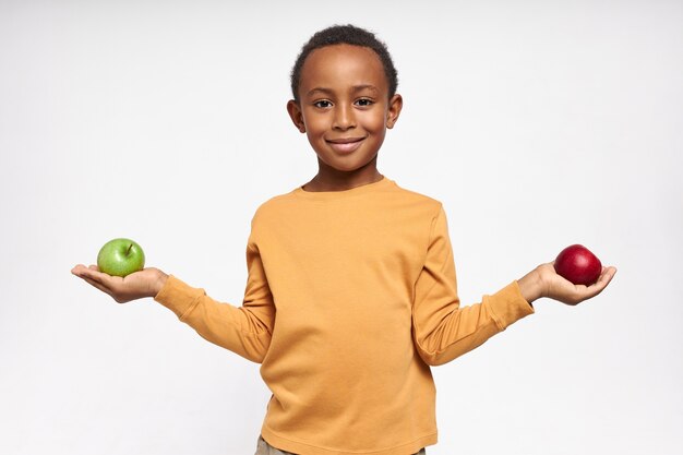 Retrato de niño negro confiado con sonrisa alegre posando aislado con manzanas verdes y rojas en sus manos