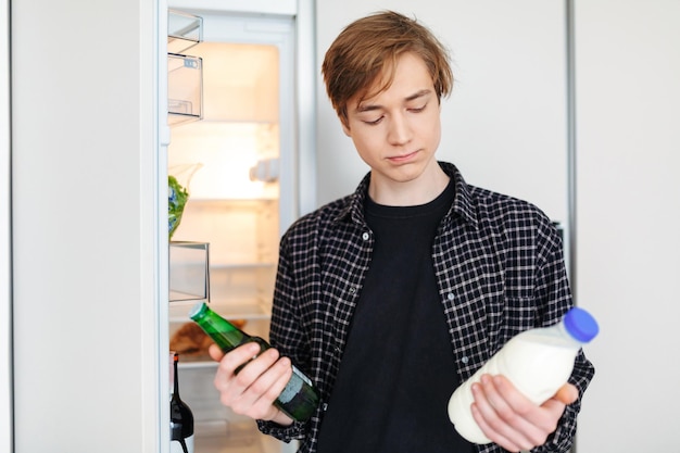 Retrato de un niño pensativo parado cerca de la nevera con cerveza y leche en las manos y decidiendo qué beber en la cocina de casa