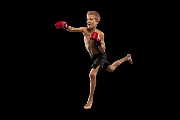 Retrato de niño pequeño en entrenamiento de uniforme protector practicando boxeo tailandés sobre fondo de estudio negro