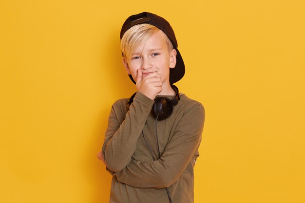 Retrato de niño pequeño con gorra negra, chico lindo posando aislado sobre amarillo, niño varón cubriendo la boca con las manos