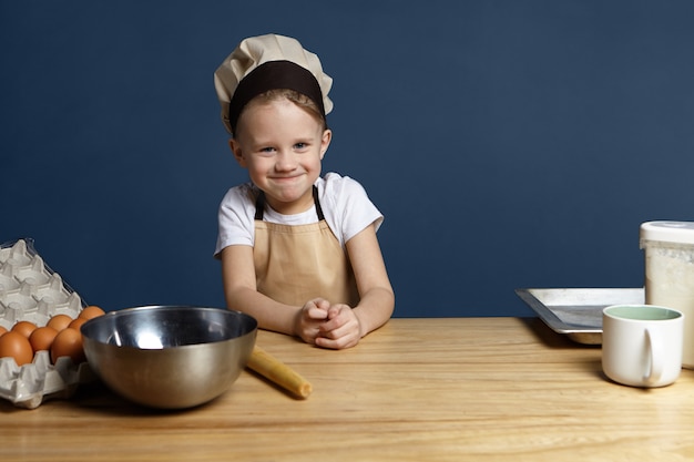 Retrato de niño pequeño lindo divertido con delantal beige y gorra de chef de pie en la cocina con taza de tazón de metal, bandeja, huevos y harina en la mesa, listo para hacer masa para pastel de pan casero o espacio de copia de pastel