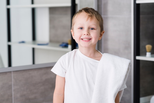 Retrato de un niño pequeño lindo sonriente con la toalla blanca sobre su hombro que se coloca en el cuarto de baño