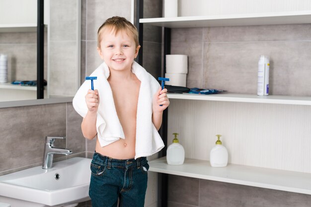 Retrato de un niño pequeño con una toalla alrededor de su cuello sosteniendo una navaja azul en las manos de pie en el baño