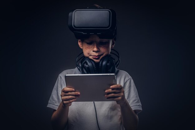 Retrato de un niño pequeño vestido con una camiseta blanca, con gafas y auriculares de realidad virtual y usando una tableta digital en un estudio, aislado en un fondo oscuro. Concepto de educación y tecnología.