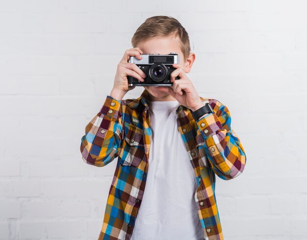 Retrato de un niño que toma una foto de una vieja cámara vintage contra una pared de ladrillo blanco