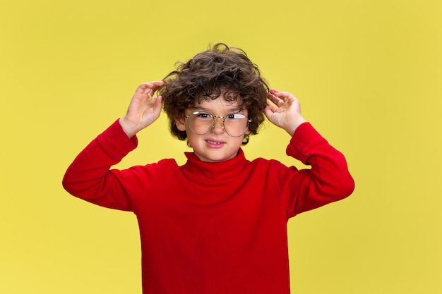 Retrato de niño rizado bastante joven en ropa roja sobre fondo amarillo de estudio. Infancia, expresión, educación, concepto de diversión.