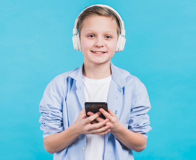 Retrato de un niño sonriente con auriculares blancos en la cabeza con teléfono inteligente en la mano contra el fondo azul