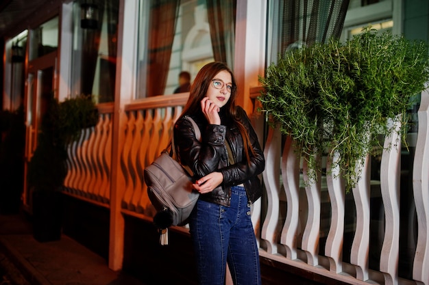 Retrato nocturno de modelo de niña con gafas, jeans y chaqueta de cuero con mochila contra las calles de la ciudad