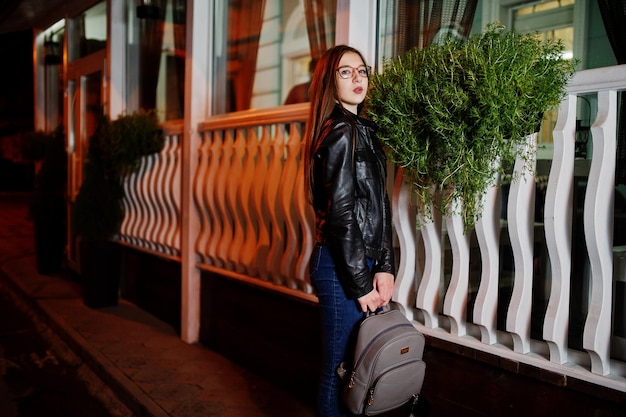 Retrato nocturno de modelo de niña con gafas, jeans y chaqueta de cuero con mochila contra las calles de la ciudad