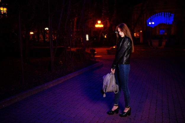 Retrato nocturno de modelo de niña con gafas, jeans y chaqueta de cuero con mochila en las manos contra la guirnalda de luces azules de la calle de la ciudad